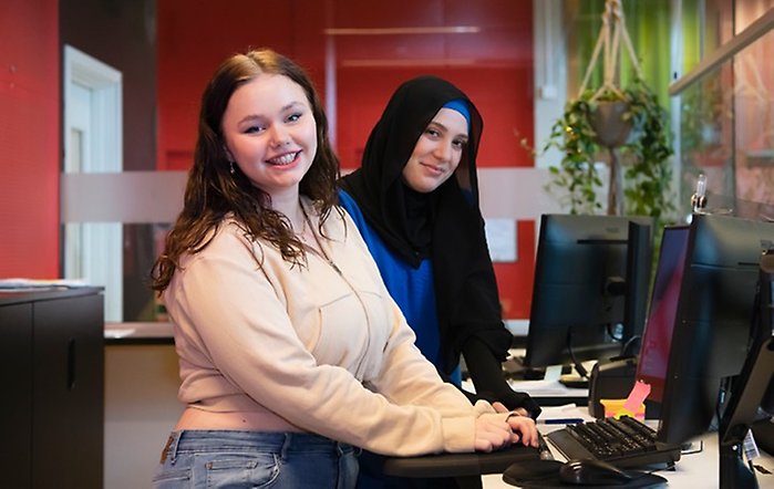 Två kvinnor står vid några datorer och tittar mot kameran och ler.
