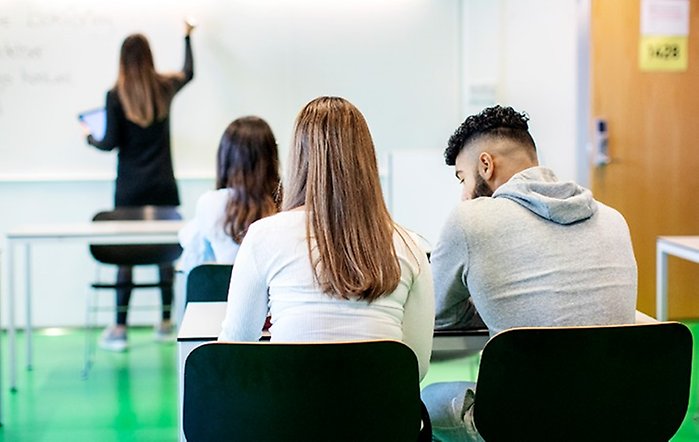 Två elever sitter i ett klassrum vända mot tavlan. I bakgrunden skriver någon på whiteboard-tavlan.