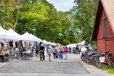 Besökare promenerar mellan tält med utställare och aktiviteter under Återbruksfestivalen på Hembygdsgården. Till höger står flera cyklar parkerade intill en röd träbyggnad.