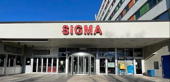 Sigma Centrums entré med den röda skylten där det står "Sigma centrum" och en klarblå himmel bakom.