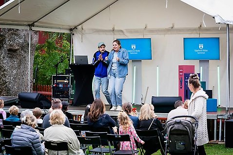 Två dramapedagoger, en man och en kvinna, från Teater & Dramaskolan uppträder på scen under Återbruksfestivalen i september. Nedanför scenen syns en publik i blandade åldrar som tittar på föreställningen